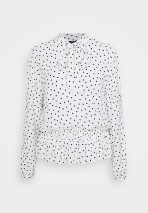 Blouse blanche à manches longues avec pois noirs, détail noué au col et ourlet volanté, présentée sur un fond uni.