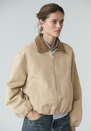 Jeune femme aux cheveux bruns attachés, portant une veste beige zippée avec un col plus foncé et un jean denim, debout devant un fond neutre.