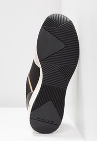 Semelle d’une basket noire et beige avec des motifs d’adhérence texturés et le logo "MICHAEL KORS" sur la partie talon.