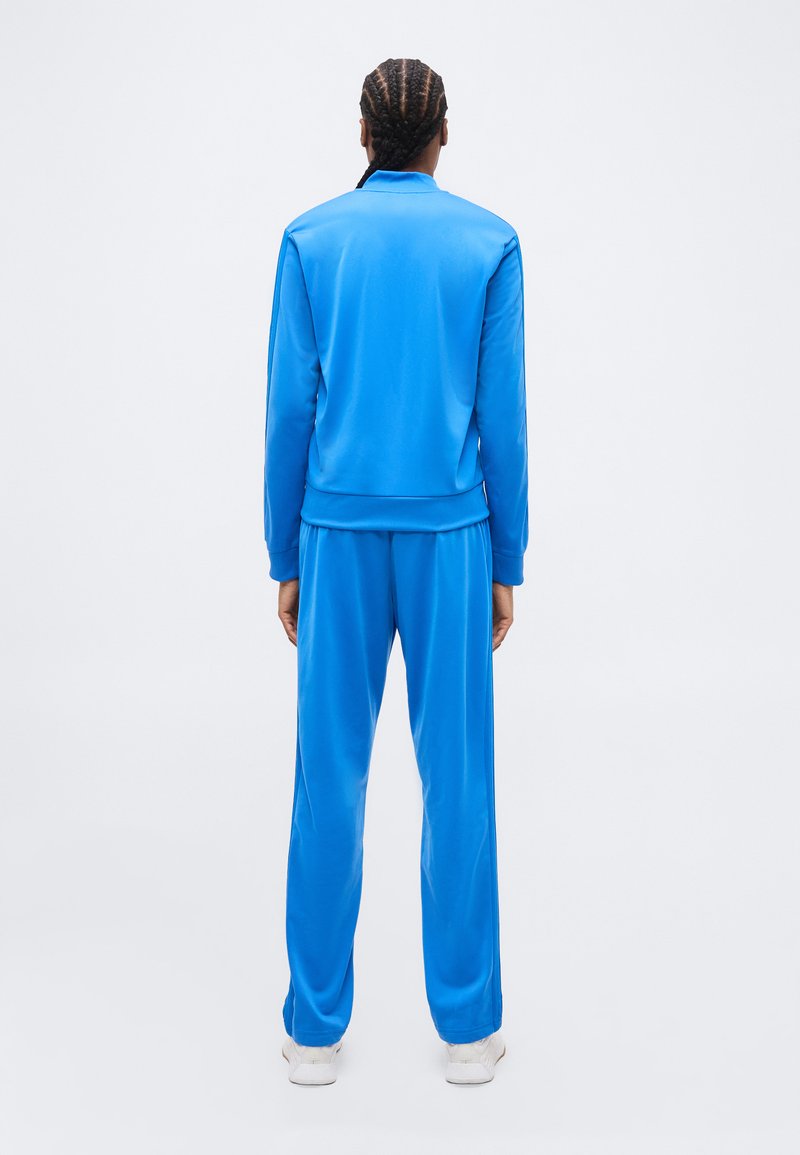 Persona con i capelli intrecciati, vista di spalle, che indossa una tuta blu brillante e scarpe da ginnastica bianche su uno sfondo neutro.