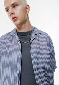 Kortærmet skjorte med knapper i blå og hvide lodrette striber, krave og broderi på brystet, lagdelt over en grå crew-neck sweater.