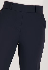 Close-up van marineblauwe slim fit broek met hoge taille en zijzakken op een lichte achtergrond.