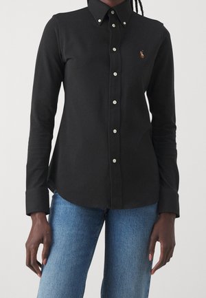 Person iført en sort knaplukket skjorte med lange ærmer med et lille broderet logo på brystet og lyseblå jeans mod en ensfarvet baggrund.