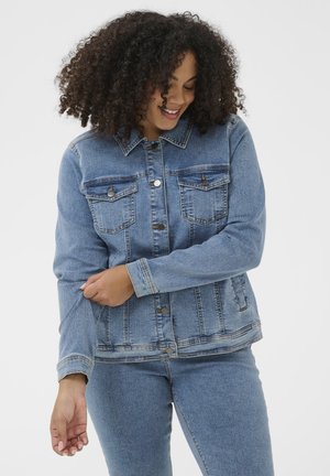 Frau mit lockigem Haar, die eine hellblaue Jeansjacke und Jeans trägt, ihren Ärmel hochkrempelt, lächelt und nach unten schaut.