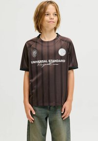 Jersey marrón con rayas que presenta un cuello redondo, mangas cortas y el texto "UNIVERSAL STANDARD" con un logotipo circular en el lado izquierdo del pecho.