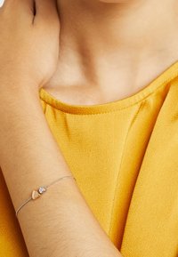 Bras reposant près de l'épaule, portant un délicat bracelet en argent avec de petits charms en forme de cœur et de cristal, sur un haut en tissu texturé jaune.