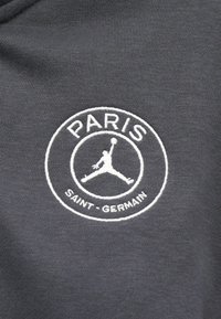 Grijze stof met een cirkelvormig geborduurd logo. De tekst bevat "PARIJS" boven en "SAINT-GERMAIN" onder, met een silhouet van een basketbal in het midden.