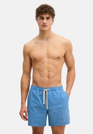 Jonge man met krullend haar, gekleed in blauwe zwembroek met witte trekkoord, staand tegen een effen lichte achtergrond.
