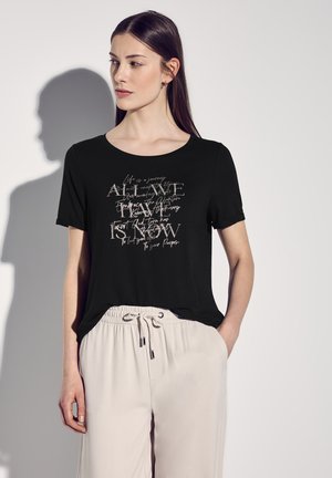 Black short-sleeve t-shirt with a light-colored motivational text graphic. Soft fabric with a relaxed fit, paired with light beige pants.