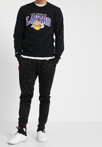 Persona che indossa una felpa nera dei Los Angeles Lakers, pantaloni jogger neri e sneakers bianche, in piedi con le mani in tasca.