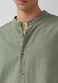 Lichtgroene blouse met een mandarinakraag, zachte textuur en lichtgrijze knopen, die een schoon en minimalistisch ontwerp laat zien.