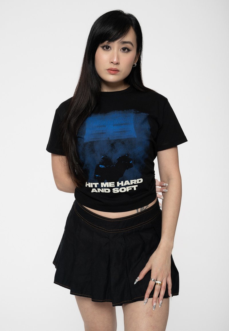 Black cropped t-shirt with blue graphic design and white text reading "HIT ME HARD AND SOFT." Paired with a black pleated skirt.