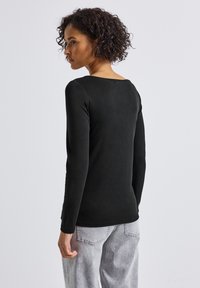 Schwarzes Langarm-Top mit rundem Ausschnitt, aus einem weichen Material, in figurbetonter Form mit dezenten Nähten. Kombiniert mit hellgrauen Hosen.