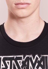 T-shirt in cotone nero con collo a costine, che presenta una grande grafica bianca, caratterizzata da un lettering audace e dettagli invecchiati.