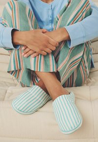 Valged ja helesinised triibulised sussid, mis on valmistatud pehmest kangast, koos roheliste ja mitmevärviliste triibuliste vabade pajama pükstega, tekstuurse beeži tausta ees.