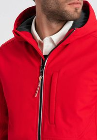 Giacca rossa con cappuccio, chiusura con zip, fodera grigia, tasca frontale e un tessuto liscio e strutturato. Presenta una cerniera nera con un tirante.