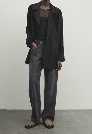 Personne portant un blazer noir, un haut noir transparent, un jean gris foncé ample et des sandales ouvertes couleur beige, debout contre un mur clair uni.