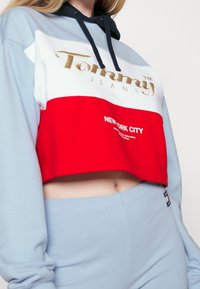 Beskuren hoodie i ljusblått med en röd och vit panel som har "Tommy Jeans" i guld och "NEW YORK CITY" tryckt nedan i vitt.