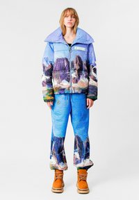 Chaqueta tipo pluma con un estampado de paisaje montañoso en azul y púrpura, combinada con pantalones de pierna ancha a juego y botas naranjas.