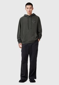 Donkergroene hoodie met een voorzak, koorden en logo-print. Gepaard met losse zwarte broek en zwarte instap schoenen.