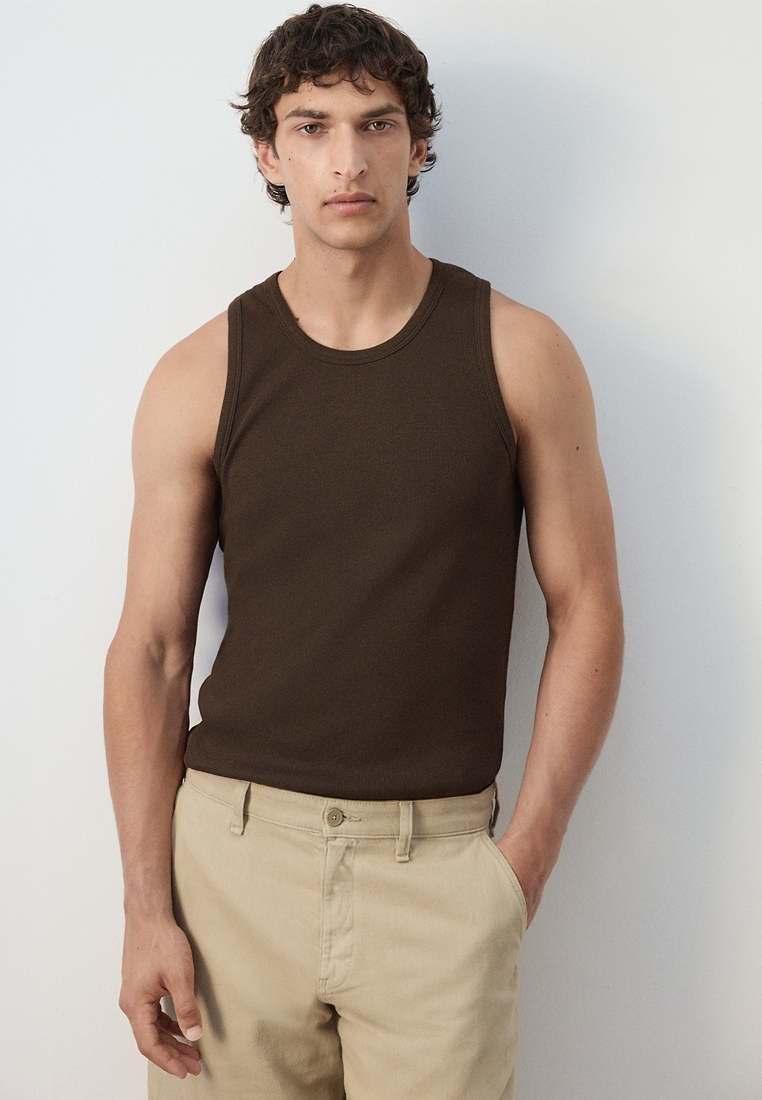 Jeune homme aux cheveux bouclés portant une chemise sans manches marron foncé et un pantalon beige, debout contre un mur gris clair.