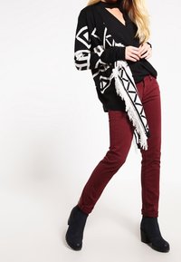 Cárdigan de patrón en blanco y negro con flecos, usado sobre una blusa negra, combinado con pantalones burdeos y botas de tobillo negras.