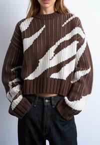 Femme portant un pull court en maille marron et blanc à motifs avec des manches longues et un jean noir taille haute, mains dans les poches.