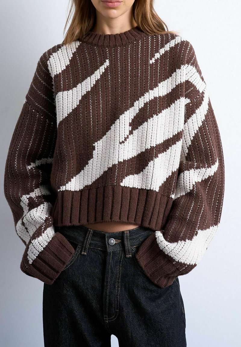 Femme portant un pull court en maille marron et blanc à motifs avec des manches longues et un jean noir taille haute, mains dans les poches.