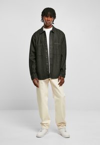 Camicia di denim nera con bottoni frontali, due taschini sul petto, indossata su una t-shirt bianca, abbinata a pantaloni color crema e sneakers bianche.