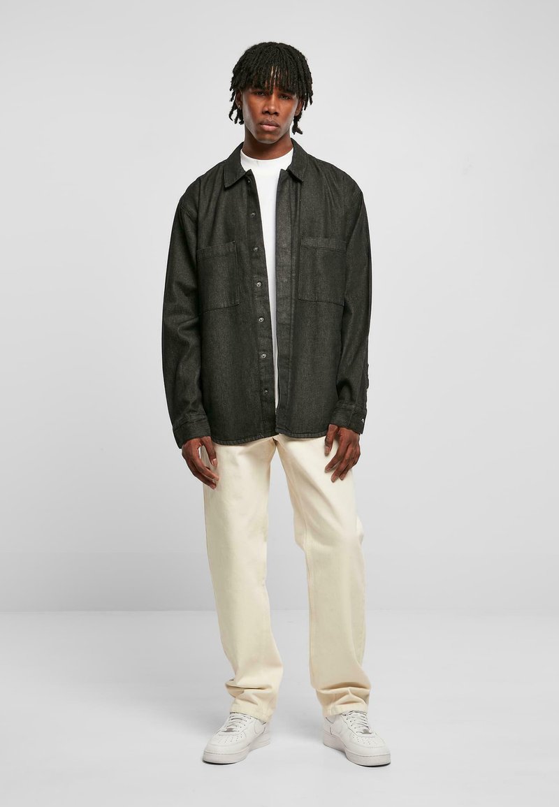 Camicia di denim nera con bottoni frontali, due taschini sul petto, indossata su una t-shirt bianca, abbinata a pantaloni color crema e sneakers bianche.