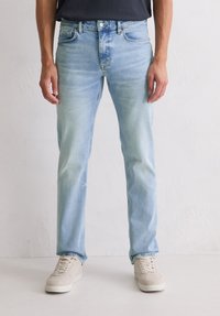 Homme portant un jean slim-fit bleu clair et des baskets beige, se tenant contre un mur blanc uni.