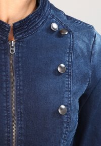 Chaqueta de mezclilla en azul oscuro con cierre de cremallera, cuello alto y cuatro botones metálicos en la parte frontal como acento. Tejido texturizado.