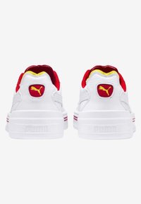 Ett par vita Puma-sneakers sedd från baksidan, med röd och gul inredning och röd Puma-logotyp på hälltappen.