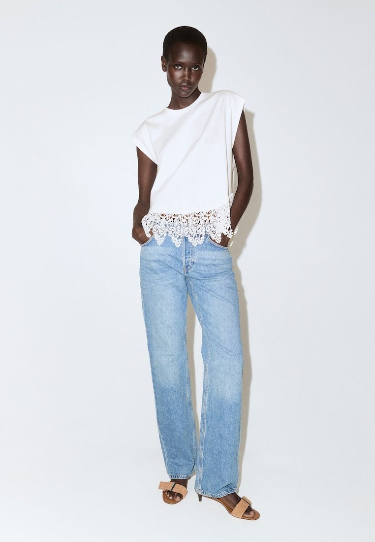 Femme à la peau foncée portant un haut blanc sans manches avec un ourlet en dentelle, un jean taille haute bleu clair et des talons ouverts couleur fauve, debout les mains dans les poches.