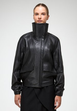 Schwarze Lederjacke mit hohem Kragen, lockerer Passform und einzigartigem Paneeldesign. Ausgestattet mit gepufften Ärmeln und strukturierter Oberfläche.