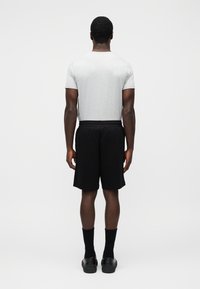 Άνδρας που στέκεται με την πλάτη στραμμένη, φορώντας ανοιχτό γκρι μπλουζάκι, μαύρο σορτς, μαύρες κάλτσες και μαύρα αθλητικά παπούτσια πάνω σε απλό φόντο.