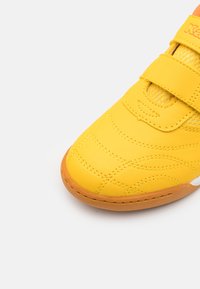 Żółty but sportowy z teksturowaną syntetyczną cholewką, przeszywaną wzorem oraz z zapięciem na rzep. Okrągły czubek i pomarańczowa gumowa podeszwa.