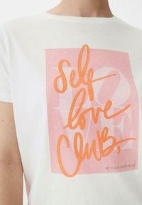 Weißes Baumwoll-T-Shirt mit einem pinken quadratischen Grafikdesign und orangefarbener Schreibschrift, die "Self love Club" liest, sowie dem Slogan "Sei deine eigene Muse."