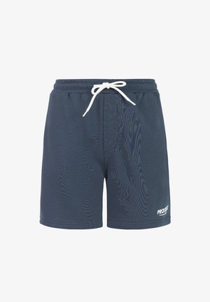 Niño rubio de pie con las manos en los bolsillos, vistiendo una camiseta beige con la inscripción "PROTEST&CO", pantalones cortos azul marino, calcetines blancos y zapatillas grises sobre un fondo blanco.