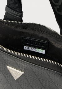 Czarna pikowana torebka na ramię z srebrnym trójkątnym logo, zamkiem błyskawicznym i wzorzystym wnętrzem z widoczną metką "GUESS".