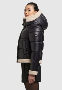 Čierna puffa bunda s plyšovým béžovým golierom a spodným lemom. Obsahuje zips, bočné vrecká a nášivku s logom na rukáve. Teplý a kompaktný dizajn.