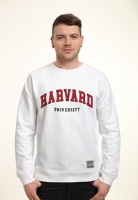 Bílá mikina s potiskem "HARVARD" a "UNIVERSITY" v červené a černé barvě. Obsahuje žebrované manžety a štítek na dolním lemu. Hladký povrch.