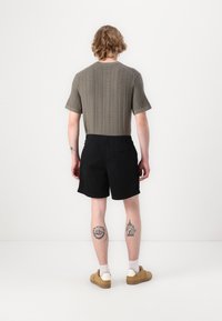 Olivenfarvet ribbet strikbluse med korte ærmer, båret med sorte shorts med elastisk talje, lette strømper og brune sko. Viser synlige tatoveringer på benene.