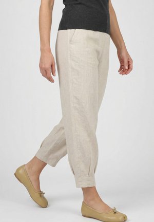 Vrouw met beige cropped broek met geplooide enkels, een zwarte top en beige ballerina’s die loopt tegen een effen achtergrond.