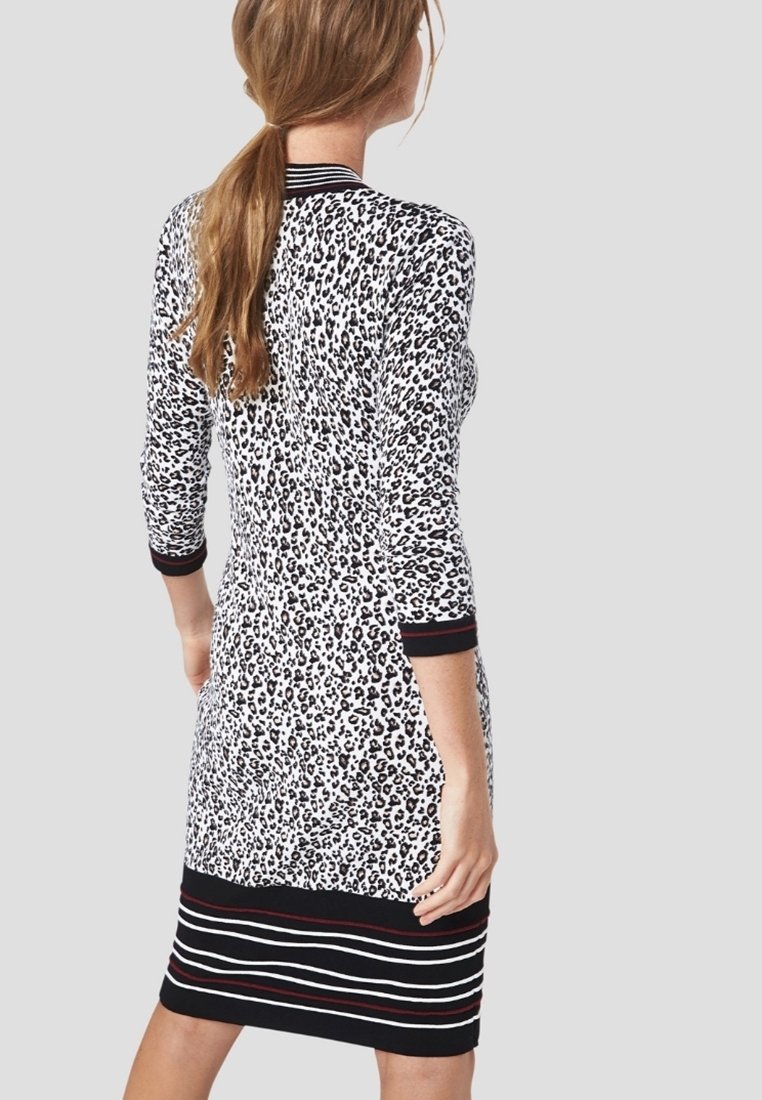 Mujer con cabello largo en una coleta, vestida con un vestido de estampado de leopardo blanco y negro, con dobladillos y puños a rayas negras y rojas, de espaldas.