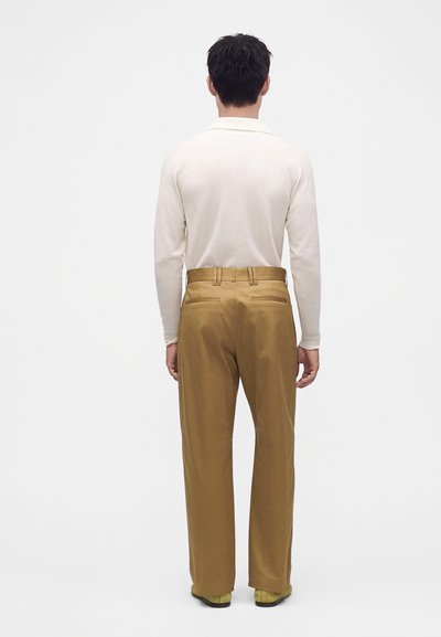 Homme debout de dos, portant une chemise blanche à manches longues et un pantalon marron taille haute droit avec des chaussures jaunes.