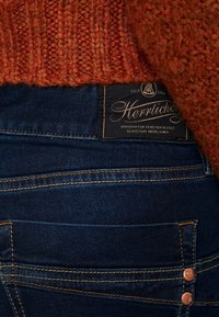 Mörkblå jeans med en strukturerad orange stickad tröja som delvis täcker dem. Funktioner inkluderar en märkesetikett och kopparfärgade knappdetaljer.