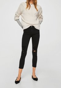 Suéter de punto beige claro con escote en V y patrón texturizado, combinado con jeans skinny negros desgastados y zapatos planos negros.