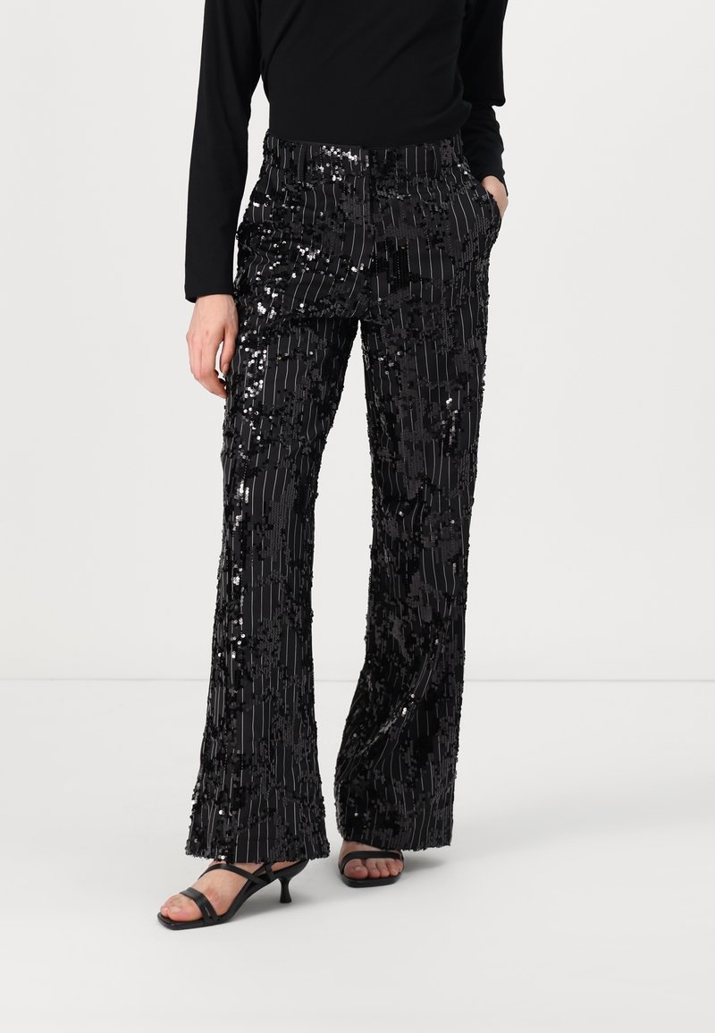 Pantalon flare noir à sequins avec des rayures verticales, porté avec un top noir à manches longues et des talons ouverts noirs. Mains dans les poches.