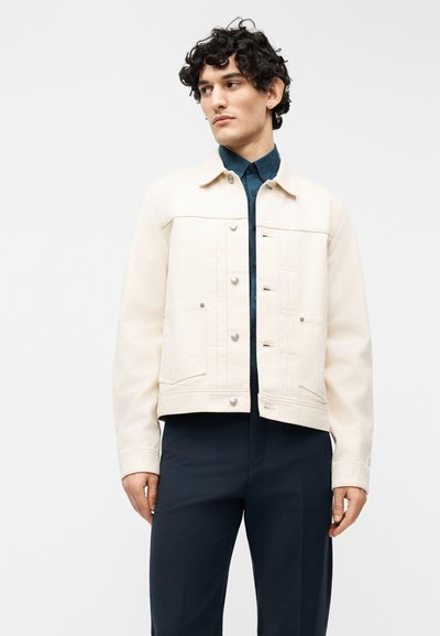 Νεαρός άνδρας με σγουρά μαλλιά που φοράει ένα κρεμ σακάκι με κουμπιά πάνω από ένα σκούρο μπλε πουκάμισο και παντελόνι που ταιριάζει, κοιτάζοντας προς τα πλάγια μπροστά σε απλό φόντο.